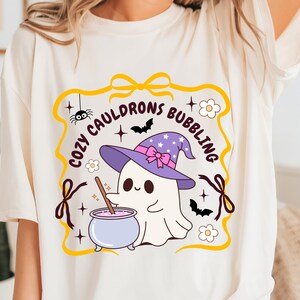 Pode incluir: Camiseta creme com um gráfico temático de Halloween. O design inclui um fantasma de desenho animado usando um chapéu de bruxa roxo com um laço rosa, mexendo em um caldeirão borbulhante. O texto "COZY CAULDRONS BUBBLING" está arqueado ao redor da imagem.