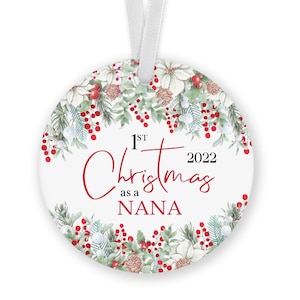May include: White round Christmas ornament with a green and red floral wreath design. The ornament has the text "1st Christmas as a Nana 2022" in red and black lettering.