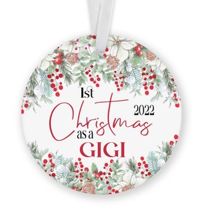May include: White round Christmas ornament with a red and green floral wreath design. The ornament says "1st Christmas as a GIGI 2022".