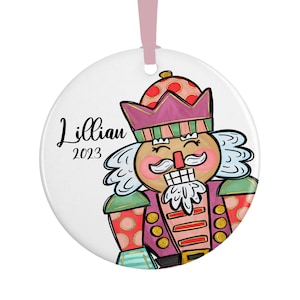 May include: A round, white Christmas ornament with a colorful illustration of a nutcracker. The nutcracker is wearing a purple hat with a red and white polka dot pattern, a green and white striped collar, and a pink and gold striped jacket. The ornament also includes the text "Lillian 2023".