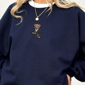 May include: Navy blue sweatshirt with a white crew neck collar. The sweatshirt features an embroidered bear wearing glasses, a green sweater, and holding a book and a mug. A gold necklace with a pendant is visible.