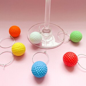 May include: A collection of colorful golf ball-shaped wine charms. The charms are in various colors: orange, yellow, white, blue, green, and red. Each charm has a silver hoop for attaching to a wine glass. The background is a solid pink.