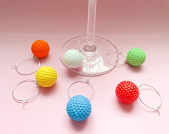 May include: A collection of colorful golf ball-shaped wine charms. The charms are in various colors: orange, yellow, white, blue, green, and red. Each charm has a silver hoop for attaching to a wine glass. The background is a solid pink.