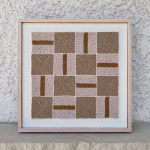 Könnte beinhalten: Ein gerahmtes Kunstwerk mit einem geometrischen Muster aus braunen und rosafarbenen Quadraten. Die Quadrate sind mit einem strukturierten, geschlungenen Stoff gefüllt.