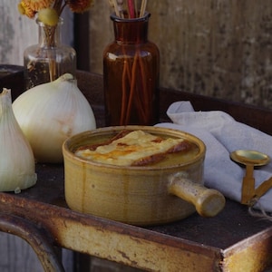 Peut inclure: Un bol de soupe à l'oignon gratinée avec du fromage fondu et du pain grillé, servi dans un bol en céramique de couleur beige rustique avec une poignée. La soupe est sur un plateau métallique avec des oignons, une cuillère et des bouteilles décoratives.