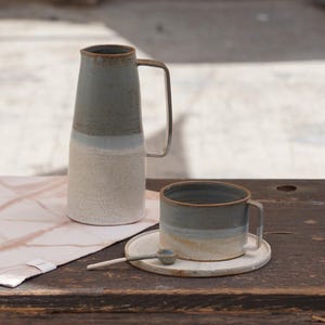 Puede incluir: Un juego de jarra y taza de cerámica con cuchara y platillo a juego. La jarra tiene un diseño alto y delgado con un asa de metal. La taza, el platillo y la cuchara son de un color similar. El juego es en tonos grises, azules y beige.