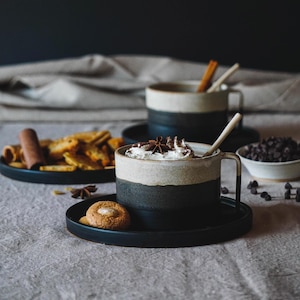 Puede incluir: Dos tazas de cerámica en crema y gris oscuro, llenas de una bebida y cubiertas con crema batida y especias. También hay un plato de galletas, un cuenco de chispas de chocolate y palitos de canela, lo que sugiere un ambiente acogedor.