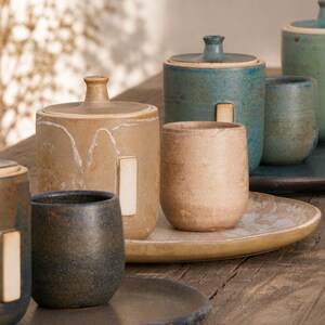 Puede incluir: Una colección de utensilios de cocina de cerámica que incluye botes con tapa y tazas en tonos tierra de beige, azul y gris oscuro. Cada pieza presenta un acabado mate suave con una pequeña pestaña rectangular en el lateral de los botes.