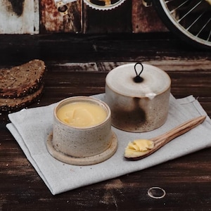 May include: A beige ceramic butter dish with a lid and a wooden spoon, filled with yellow butter. The dish has a speckled design and sits on a grey linen cloth. Slices of bread are in the background.