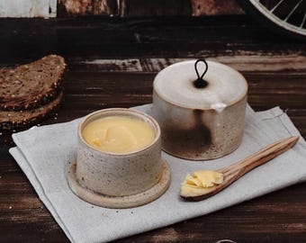 May include: A ceramic butter dish with a lid and a wooden spoon, filled with yellow butter. The dish is beige with a speckled design, and sits on a gray linen cloth. Slices of bread are in the background.