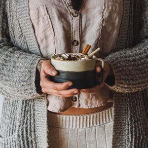 Puede incluir: Una persona sostiene una taza de cerámica de dos tonos llena de una bebida caliente, cubierta con crema batida, virutas de chocolate y una ramita de canela. La taza es negra y beige. La persona lleva una rebeca gris y una camisa rosa.