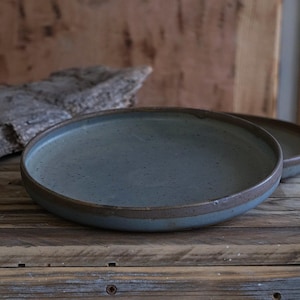 Puede incluir: Dos platos de cerámica con una superficie moteada azul-verdosa mate y un borde marrón oscuro. Los platos están sobre una superficie de madera. Adecuados para servir alimentos.