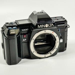 May include: A black Minolta 7000 Maxxum AF 35mm SLR camera body. The front features a circular lens mount with a silver metal ring, a red dot alignment marker, and the text Minolta, Maxxum, 7000, and AF printed in white on the black plastic casing.