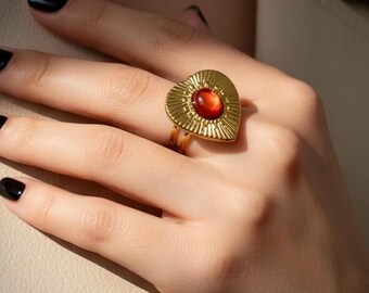 May include: A gold-colored ring with a heart-shaped design and a central orange-red stone. The ring has a textured, sunburst-like pattern around the stone. The ring is on a person's finger, with black nail polish.