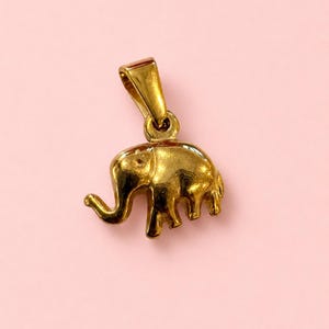May include: A gold-colored elephant pendant with a loop for a chain. The elephant has a trunk, tusks, and four legs. The pendant is likely made of metal and is displayed against a pink background. This jewelry item is suitable for necklaces.