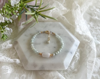 May include: A delicate bracelet made with light green beads and a larger, white oval-shaped bead. The bracelet has a gold clasp and a small gold charm.