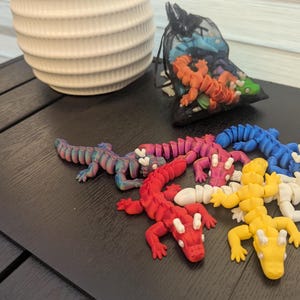 May include: Six colourful plastic toy dragons, each with a different colour: red, pink, blue, white, yellow, and iridescent. The dragons are posed in a variety of positions on a black surface.