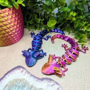 May include: Two 3D printed lizard figurines, one is purple and blue, the other is pink and orange. The lizards are posed in a curled up position.