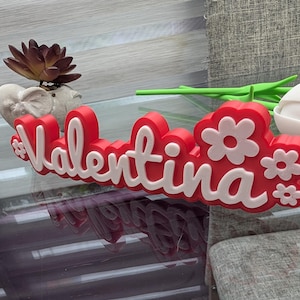 May include: A red and white decorative sign that spells out "Valentina" in a cursive font. The sign is decorated with white flower shapes. A small succulent plant in a heart-shaped pot and a white tulip are also visible.