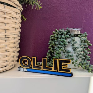 May include: A black wooden name sign with the name "OLLIE" in yellow letters. The sign has a blue and yellow lightsaber underneath it.