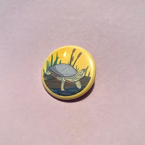 Puede incluir: Un botón amarillo con una ilustración de dibujos animados de una tortuga nadando en un estanque con juncos verdes. La tortuga tiene un caparazón marrón con la parte inferior gris y patas naranjas.