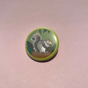 May include: A round button with a green border featuring a gray squirrel eating a nut. The squirrel is depicted on a brown log with green leaves in the background. The button is set against a pink background.
