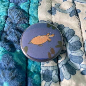 May include: A blue button with a brown turtle swimming in a blue pond with green reeds. The turtle has a light brown shell and a dark brown head and legs.