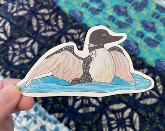 May include: A sticker of a loon with a starry pattern on its back, swimming in a blue lake. The loon has a black head and neck with a red eye.