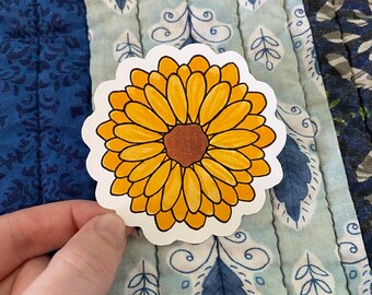 May include: A die-cut sticker featuring a yellow sunflower with a brown center. The flower has detailed petals and is set against a white background. The sticker is held against a blue patterned quilt.