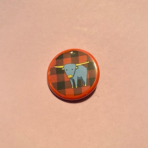 Może przedstawiać: Okrągła przypinka z czerwono-czarnym wzorem w kratę. Na środku znajduje się ilustracja niebieskiej krowy szkockiej z żółtymi rogami. Dekoracyjny dodatek.