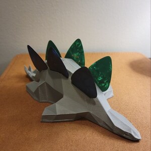 May include: A gray 3D printed dinosaur-shaped guitar pick holder with five guitar picks inserted. The picks are green and black.