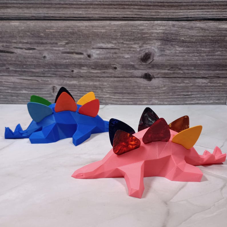 May include: Two colorful dinosaur-shaped guitar pick holders. The blue dinosaur has a red, orange, yellow, and green pick on its back. The pink dinosaur has a black, red, orange, and yellow pick on its back.