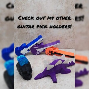 May include: A collection of guitar pick holders, including a purple dinosaur, a black skull with a blue mohawk, and three guitar-shaped holders in blue, purple, and orange.