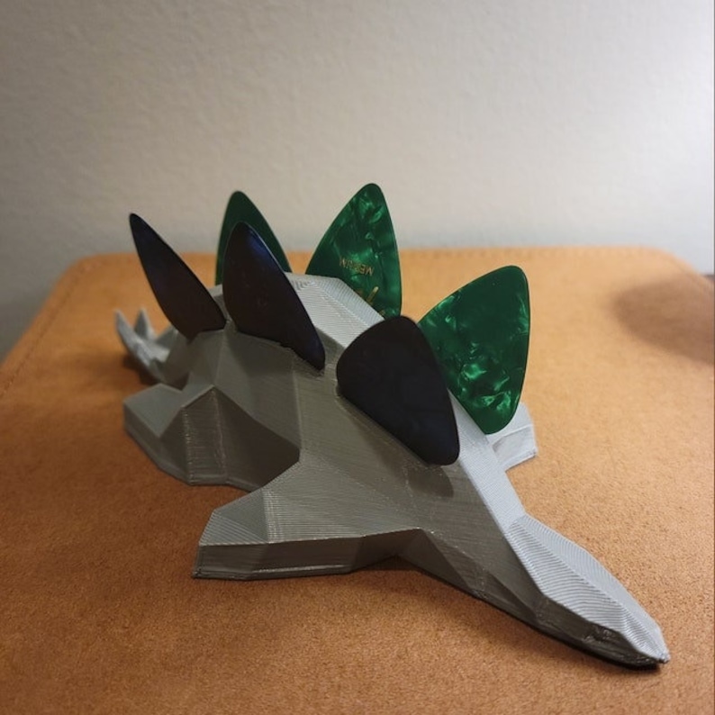 May include: A gray 3D printed dinosaur-shaped guitar pick holder with five guitar picks inserted. The picks are green and black.