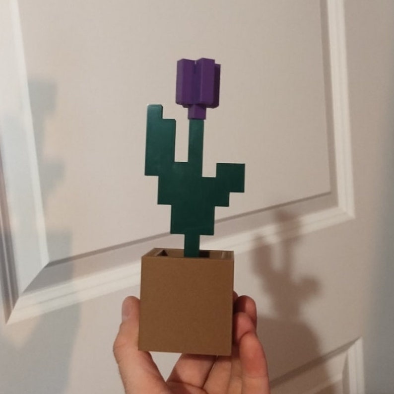 May include: A 3D printed Minecraft flower pot with a purple flower and green leaves. The pot is brown and the flower is in a simple, blocky style.