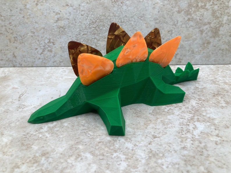 May include: Green 3D printed dinosaur shaped guitar pick holder with orange and brown guitar picks.