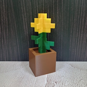 Pode incluir: Um vaso de flores Minecraft impresso em 3D com uma flor amarela e um caule verde. O vaso é marrom e tem formato quadrado.
