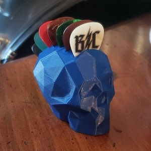 May include: A blue 3D printed skull shaped guitar pick holder with a variety of guitar picks inserted into the top. The top pick has the text "G&C" on it.