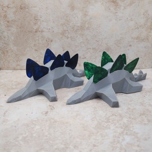 May include: Two gray dinosaur-shaped guitar pick holders. One holder has blue picks and the other has green picks.