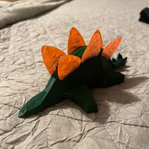 May include: A green 3D printed dinosaur with orange guitar picks on its back. The picks are labeled "Jim Dunlop" and "Medium".