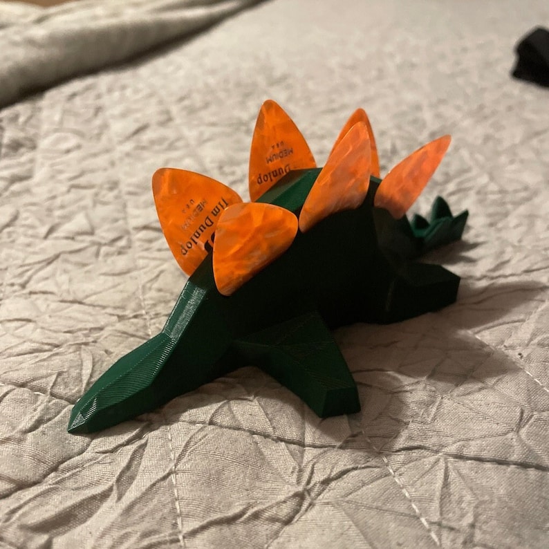 May include: A green 3D printed dinosaur with orange guitar picks on its back. The picks are labeled "Jim Dunlop" and "Medium".