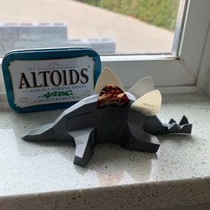 May include: A gray 3D printed dinosaur-shaped guitar pick holder with three guitar picks inserted. The picks are white and brown with the text "medium" and "genuine" printed on them. A blue and white Altoids tin is in the background.