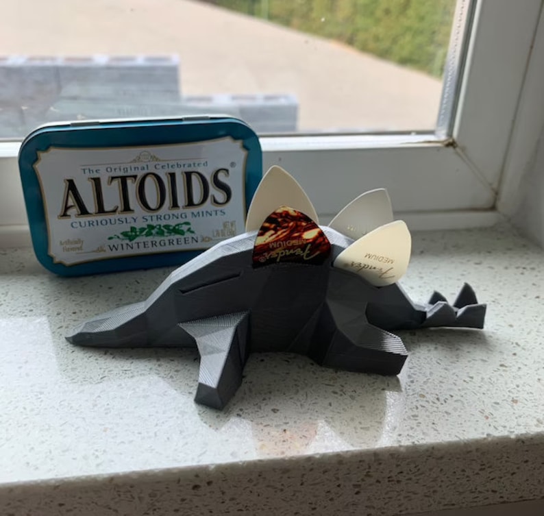 May include: A gray 3D printed dinosaur-shaped guitar pick holder with three guitar picks inserted. The picks are white and brown with the text "medium" and "genuine" printed on them. A blue and white Altoids tin is in the background.