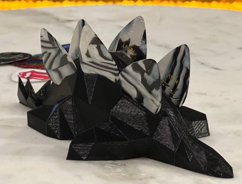 May include: A black 3D printed guitar pick holder with five guitar picks. The picks are white and black with a swirl pattern and have the word "JIMMY" written on them.