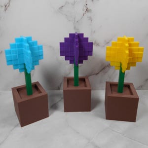Peut inclure: Trois sculptures décoratives de fleurs bleues, violettes et jaunes. Chaque fleur a un design pixélisé, une tige verte et repose dans un pot carré marron. Les fleurs sont disposées en rangée, mettant en valeur leurs couleurs vives et leur style unique.