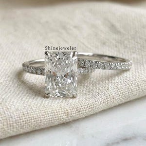 May include: A radiant-cut diamond engagement ring with a matching band. The rings are set in a silver band and rest on a neutral-colored fabric. The text "ShineJeweler" is visible on the image.