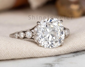 May include: A silver-toned ring featuring a large, cushion-cut center stone held by four prongs. The band is split and decorated with small, round, clear stones set in a milgrain-detailed metal frame. The text Shinejeweler is visible in the background.