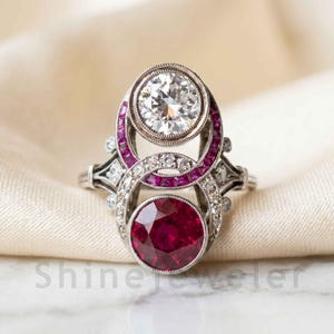 Può includere: Un anello in argento con un grande diamante bianco rotondo nella parte superiore e un grande rubino rosso rotondo nella parte inferiore. Il centro è accentuato da rubini rossi e diamanti bianchi più piccoli incastonati in una montatura traforata.