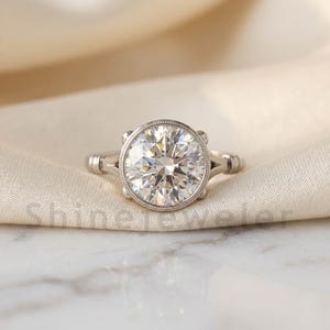 May include: A silver ring featuring a large, round-cut diamond. The diamond is set within a bezel and is the central feature. The ring is presented on a white, textured surface.