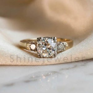 May include: A vintage-style engagement ring with a large, sparkling, round-cut diamond as the centerpiece. The ring has a gold band with smaller accent diamonds on each side of the main stone. The setting is a classic, elegant design.